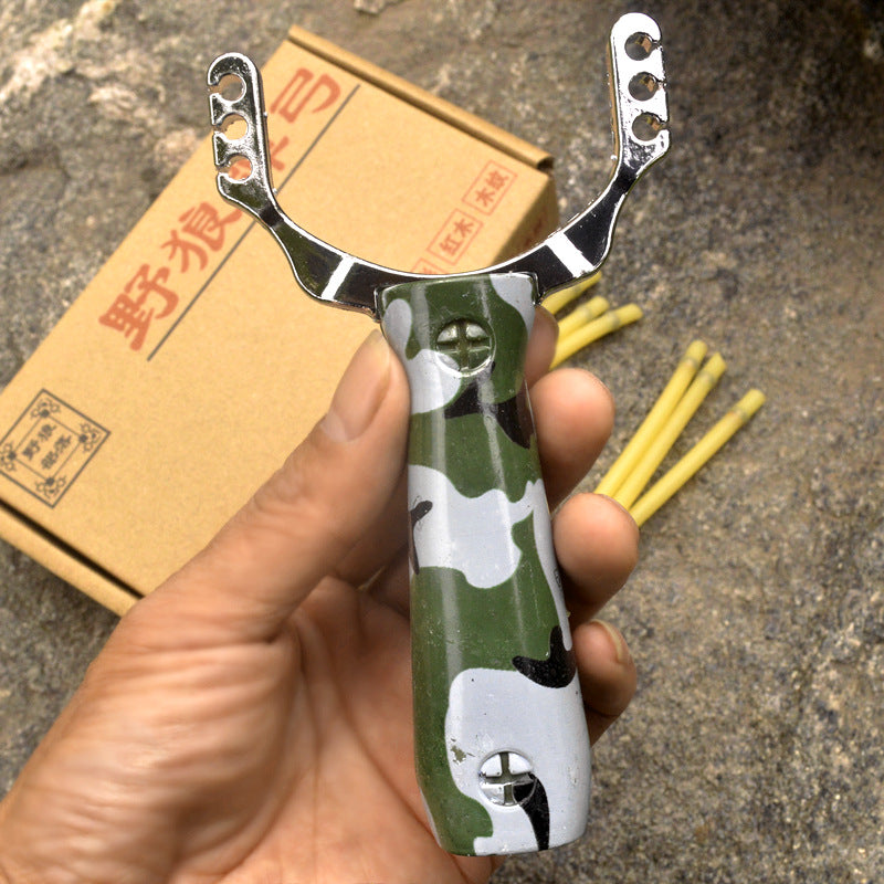 Hand holding the camouflage slingshot outdoors with the printed cardboard box and yellow band strands behind