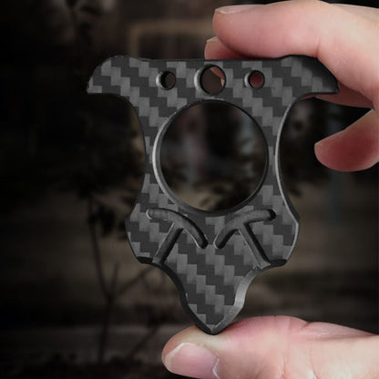 Cat-ear shaped carbon fiber knuckle with a central finger hole and three top holes, held between fingers against a dark blurred foliage backdrop.