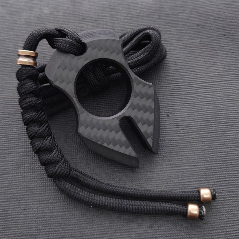 Claw-shaped carbon fiber knuckle with a two-pronged forked tip and a central finger hole, hung from a black paracord finished with copper bead caps on dark cordura.
