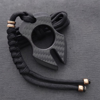 Claw-shaped carbon fiber knuckle with a two-pronged forked tip and a central finger hole, hung from a black paracord finished with copper bead caps on dark cordura.