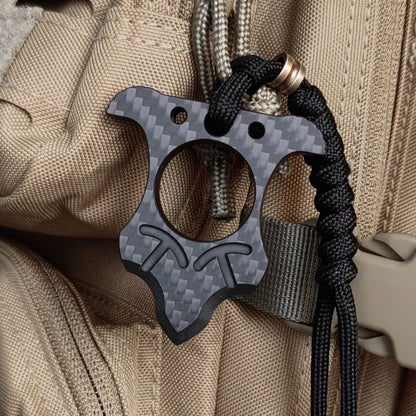 Crown-shaped carbon fiber knuckle with a central finger hole and horned top, hung from a knotted paracord with a copper bead on a tan tactical pack.