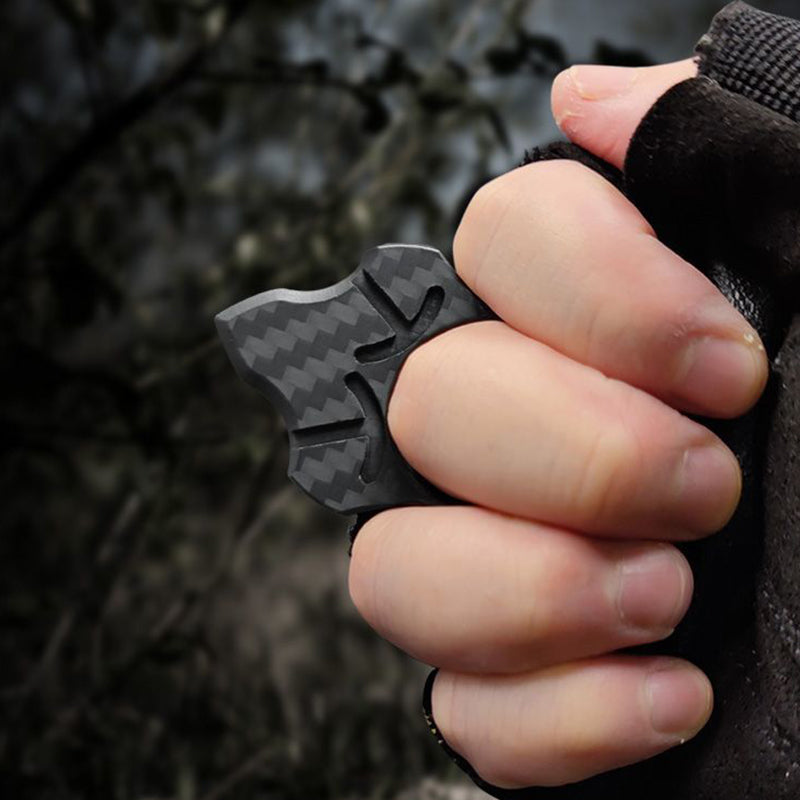 Carbon fiber single-finger knuckle worn on the index finger of a closed fist in a black fingerless glove, against a blurred woodland backdrop.