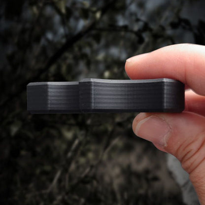 Small black carbon fiber knuckle pinched horizontally between the thumb and fingers in a blurred woodland setting.