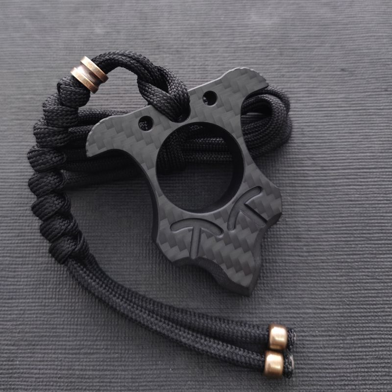 Shield-shaped carbon fiber knuckle with horned top and scalloped sides, hung from a black paracord with copper beads against a dark charcoal mat.