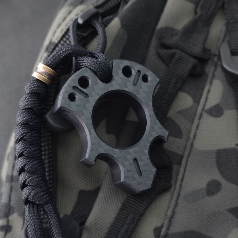 Talon-shaped carbon fiber knuckle with milled teardrop slots and a central finger hole, hung from a black paracord with a bronze bead on a camouflage backpack.