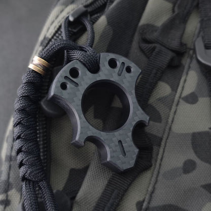 Talon-shaped carbon fiber knuckle with milled teardrop slots and a central finger hole, hung from a black paracord with a bronze bead on a camouflage backpack.