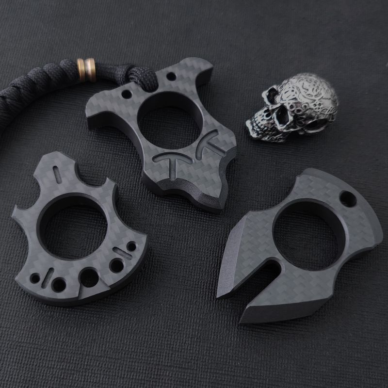 Three carbon fiber single-finger knuckle styles laid out on a black textured surface with a metal skull bead: an axe-shaped one, a bat-wing one on a paracord lanyard, and a claw-shaped one.