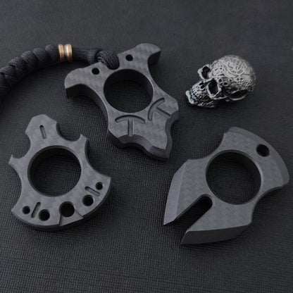 Three carbon fiber single-finger knuckle styles laid out on a black textured surface with a metal skull bead: an axe-shaped one, a bat-wing one on a paracord lanyard, and a claw-shaped one.