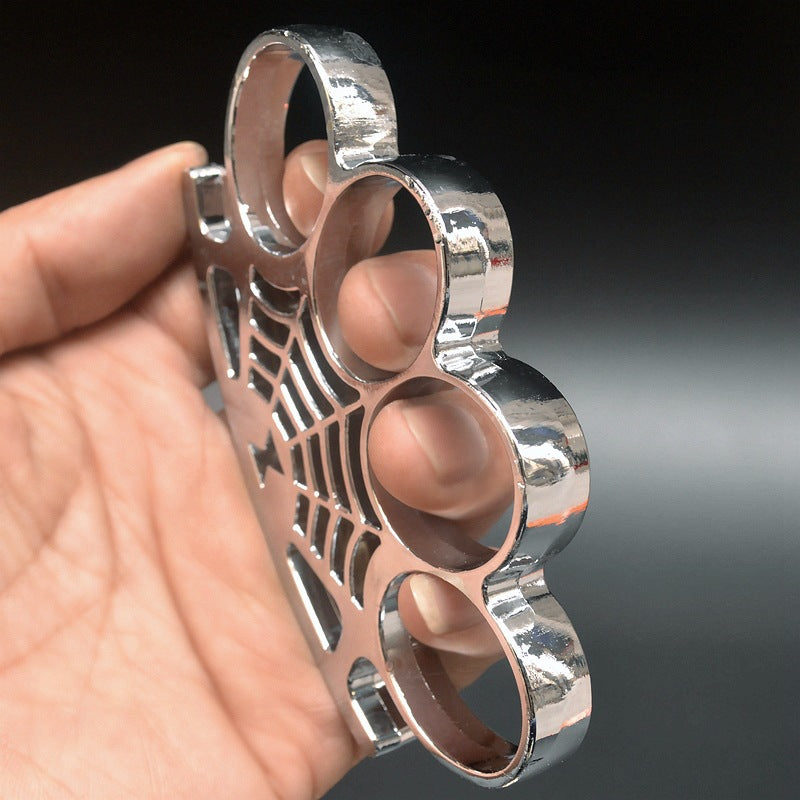 A chrome-finished knuckle duster held in a hand from a side angle, with four oval finger rings and a spider web pattern with a spider on the palm section.