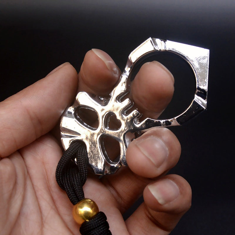 A high-polish chrome skull knuckle duster fitted over one finger against a dark background, the heart-shaped hole and eye sockets cut clean through the skull face, with a black paracord lanyard and gold bead below.