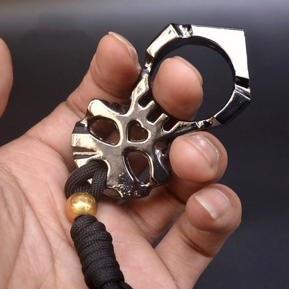 A chrome two-tone skull knuckle duster worn on one finger, the ring at the top and the skull below with its heart-shaped hole and eye sockets, a black paracord lanyard with a gold bead hanging from the base.