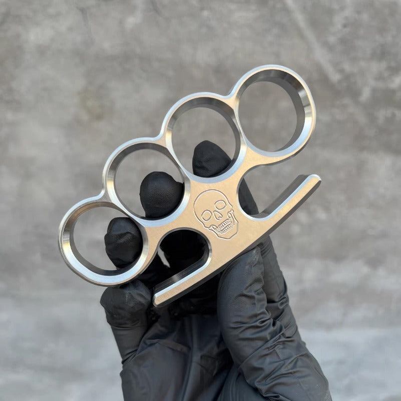 The 304 stainless steel classic knuckle duster stands upright between a black-gloved thumb and fingers.