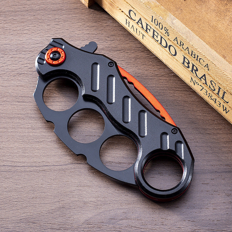 Closed black folding knuckle knife with red accents on wood next to a coffee bag
