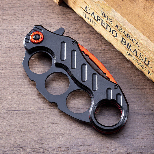 Closed black folding knuckle knife with red accents on wood next to a coffee bag
