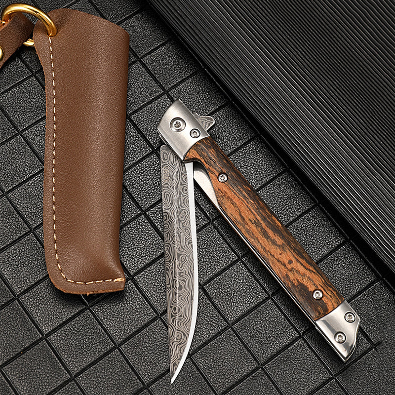 Closed folding knife with a Damascus-pattern blade folded into its wood handle, lying beside a brown leather sheath with brass ring on a dark grid-patterned mat