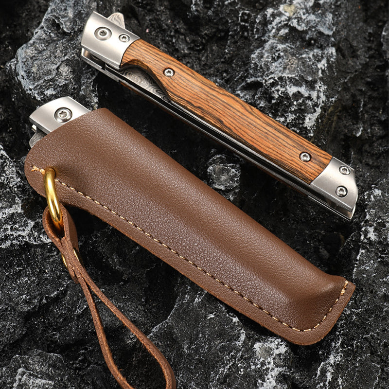Closed folding knife with striped wood handle and steel bolsters beside a stitched brown leather sheath on dark rock