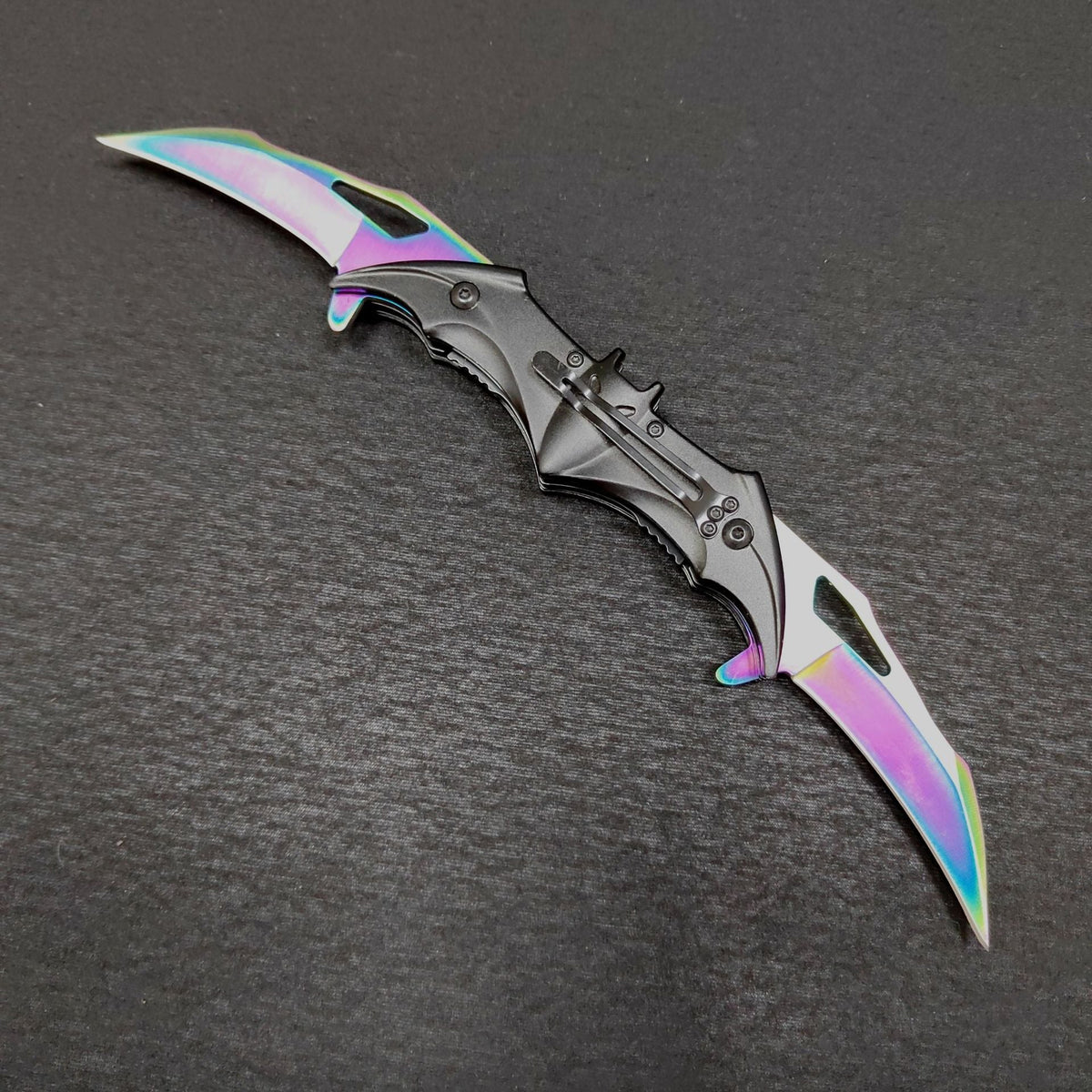 The colourful rainbow bat knife shown fully open from the clip side on a dark gray surface, two iridescent blades extended from a black bat handle with a fixed pocket clip.