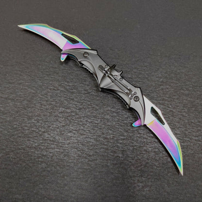 The colourful rainbow bat knife shown fully open from the clip side on a dark gray surface, two iridescent blades extended from a black bat handle with a fixed pocket clip.