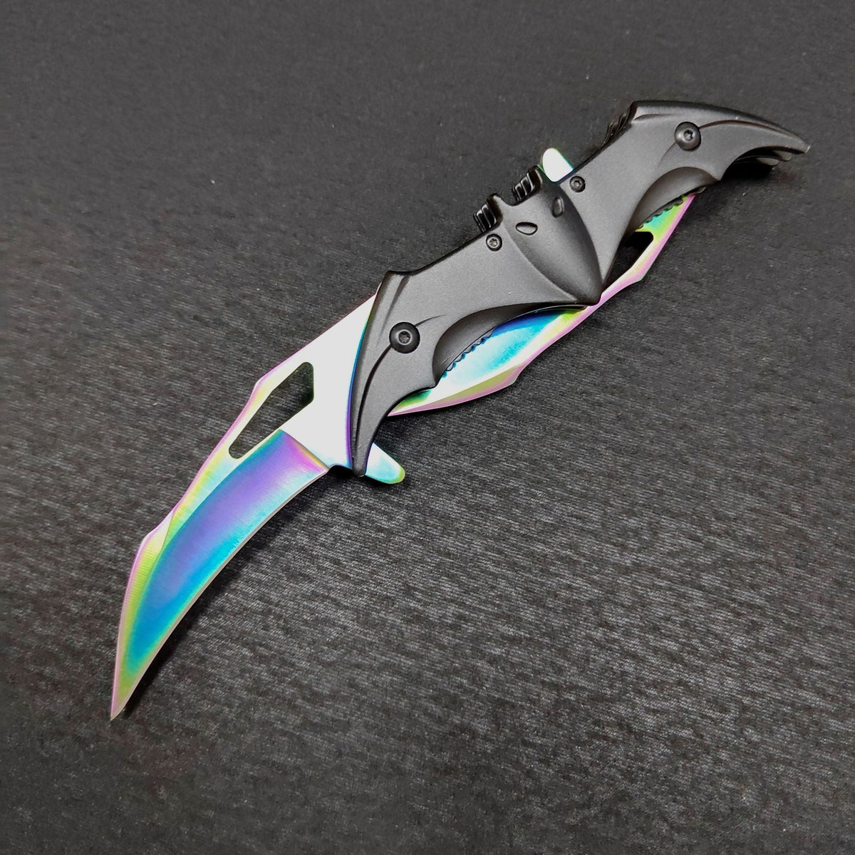 The colourful rainbow bat knife fully open on a dark gray surface, both iridescent blades extended from the black bat face handle with its ears, eyes and wing slots.