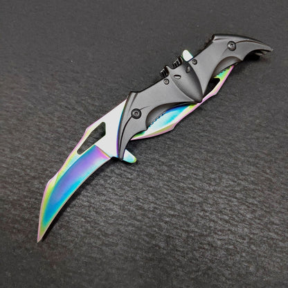 The colourful rainbow bat knife fully open on a dark gray surface, both iridescent blades extended from the black bat face handle with its ears, eyes and wing slots.