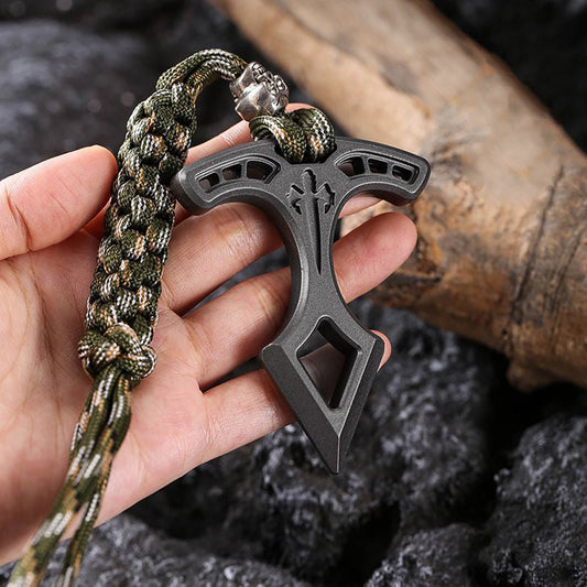 Hand holding the metal pendant by its camo paracord lanyard, showing the slotted T-bar and engraved cross against dark rock.