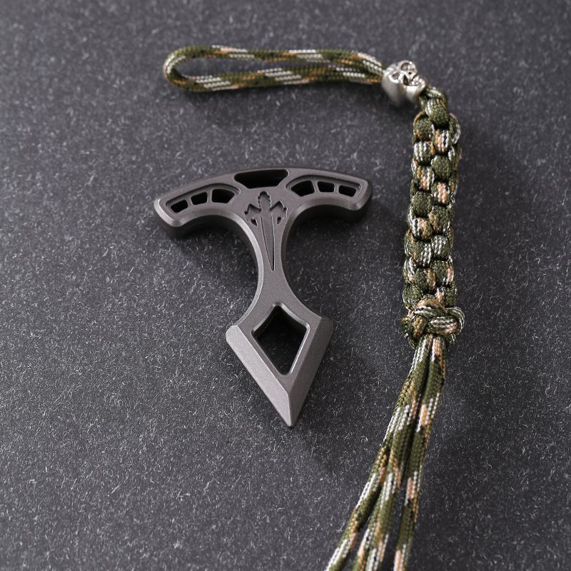 Metal pendant lying on a dark textured surface beside a camo paracord lanyard with a steel bead and frayed tassel.