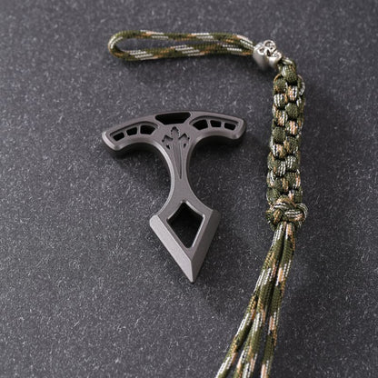 Metal pendant lying on a dark textured surface beside a camo paracord lanyard with a steel bead and frayed tassel.