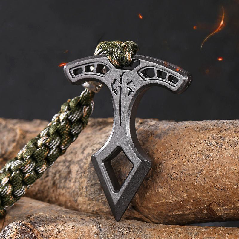Spear head EDC pendant standing upright on a log with embers in the background, showing the slotted bar and pointed tip.