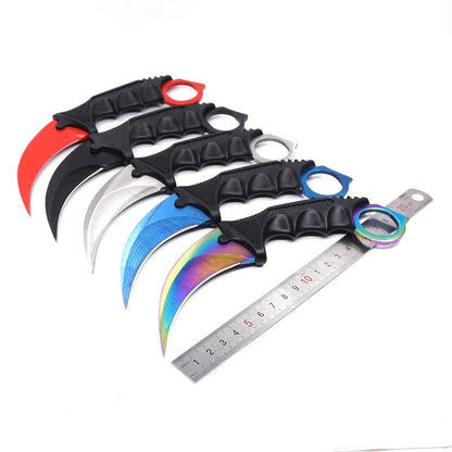 Five karambit knives with red black silver blue and rainbow blades beside a ruler