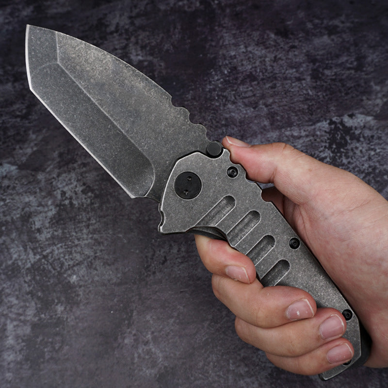 A hand grips the open folding knife against a mottled dark background, showing its full size with the stonewashed blade and the grooved stonewashed steel handle.