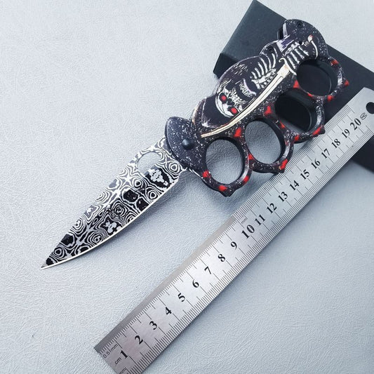 Folding EDC knife with four finger openings and black-red skeleton artwork, open beside a steel ruler showing about 22cm overall length