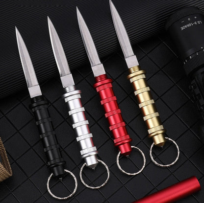 Four keychain daggers in Black, Silver, Red and Gold laid diagonally on a black grid mat, blades exposed, each with a ribbed handle and a keyring.