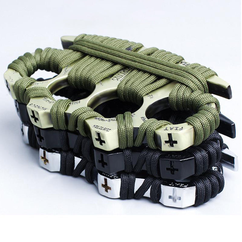 Four zinc alloy knuckle dusters stacked and overlapping on a white background, wrapped in paracord - an olive-green wrapped golden one on top and black-wrapped black, silver and bronze examples below, each with a cross motif and lettering embossed on the knuckle face.