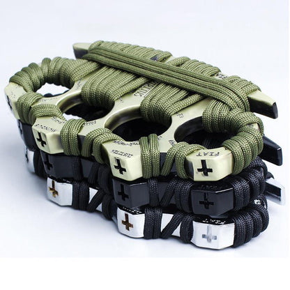 Four zinc alloy knuckle dusters stacked and overlapping on a white background, wrapped in paracord - an olive-green wrapped golden one on top and black-wrapped black, silver and bronze examples below, each with a cross motif and lettering embossed on the knuckle face.
