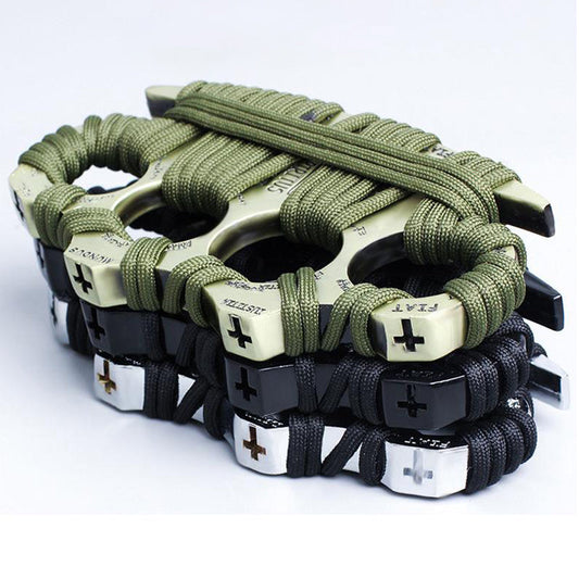 Four zinc alloy knuckle dusters stacked and overlapping on a white background, wrapped in paracord - an olive-green wrapped golden one on top and black-wrapped black, silver and bronze examples below, each with a cross motif and lettering embossed on the knuckle face.
