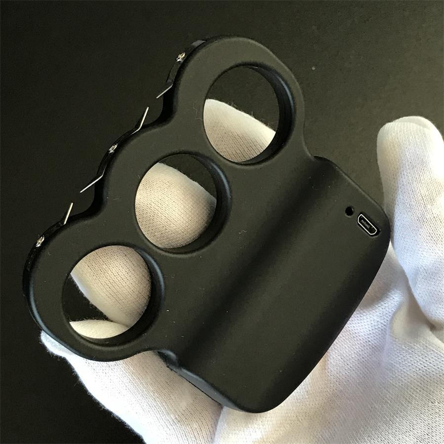 White-gloved hand holding the black three-hole stun device face up, three finger rings and pointed prongs along the upper left edge, USB port on the right