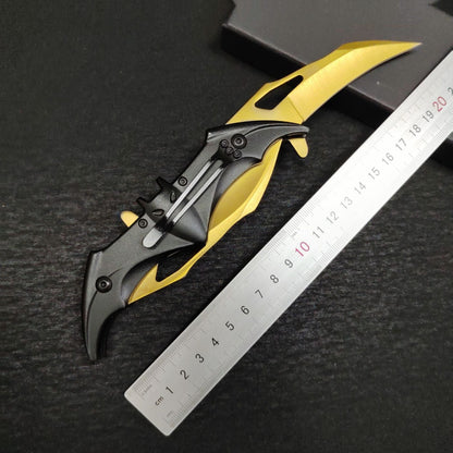 The back of the fully open gold bat knife beside a metal ruler, showing a black pocket clip with a bright steel strip set into the bat-shaped handle, with gold wing edges and slotted edges.
