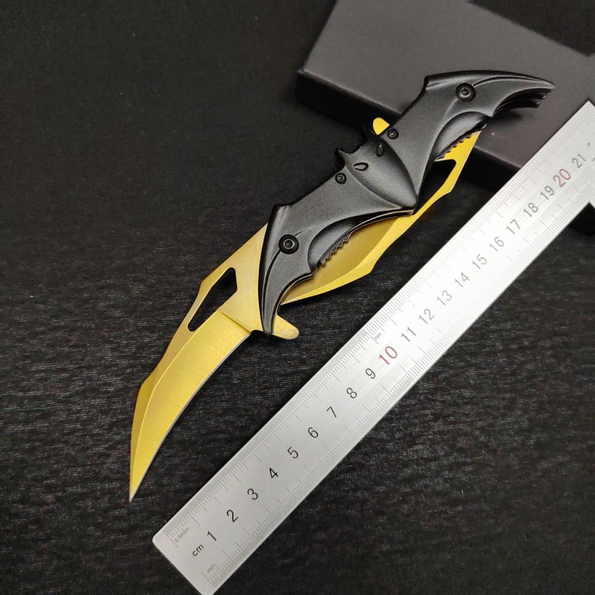 A bat-shaped folding knife with a gold blade extended beside a metal ruler for scale, its black bat handle showing ears, eyes and a serrated section, with a black box in the background.