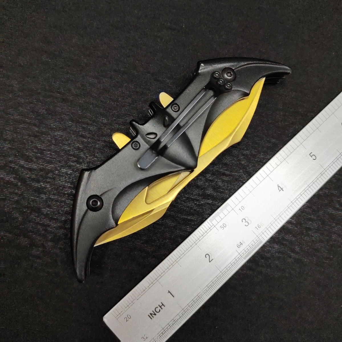 The closed gold bat knife seen from the clip side beside a dual-scale ruler, showing the black pocket clip fixed to the bat handle and the gold wing edges folded underneath.