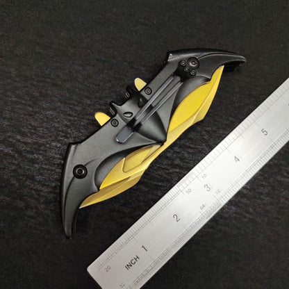 The closed gold bat knife seen from the clip side beside a dual-scale ruler, showing the black pocket clip fixed to the bat handle and the gold wing edges folded underneath.