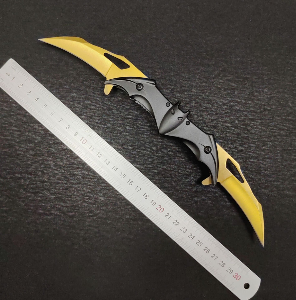 The fully open gold bat knife measured against a ruler from end to end, showing two gold blades extended from a black bat handle with ears, eyes and slotted edges.