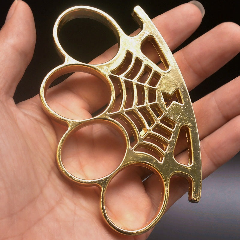 A gold four-finger knuckle duster held at an angle in the palm, with four oval finger holes and a spider-web casting running along the curved palm rest.