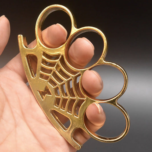 A gold four-finger knuckle duster held in a hand, with four large oval finger holes and a cast spider-web pattern with a spider across the palm rest.