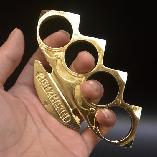 Gold knuckle duster held in hand showing four finger holes and the embossed palm plate on a dark background