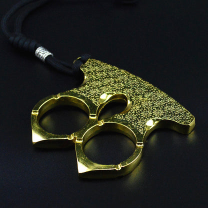 Gold two-finger knuckle with an engraved floral plate lying flat on a dark surface, braided cord and metal bead running up