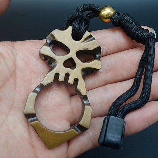 A hand holds a gold two-tone single-finger skull knuckle duster, its face showing two eye sockets and a heart-shaped hole above one large finger ring, with a black braided paracord lanyard, gold bead and black cord lock attached.