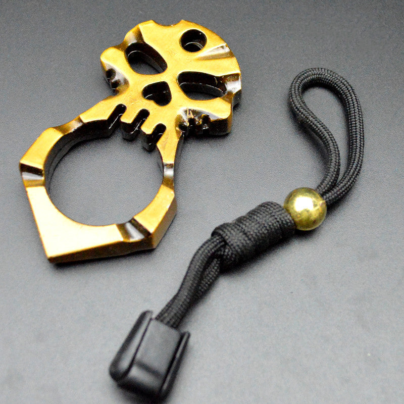 The polished gold skull knuckle duster laid beside its black paracord lanyard, gold bead and black cord lock on a dark gray surface, with the heart-shaped hole and eye sockets clearly cut through the face.