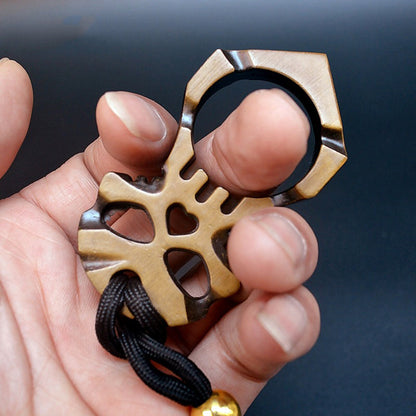 Close-up of the gold two-tone skull knuckle duster worn on one finger, the large ring at the top and the skull face below showing two eye sockets, a heart-shaped hole and teeth cut-outs, with a black paracord lanyard through the base.