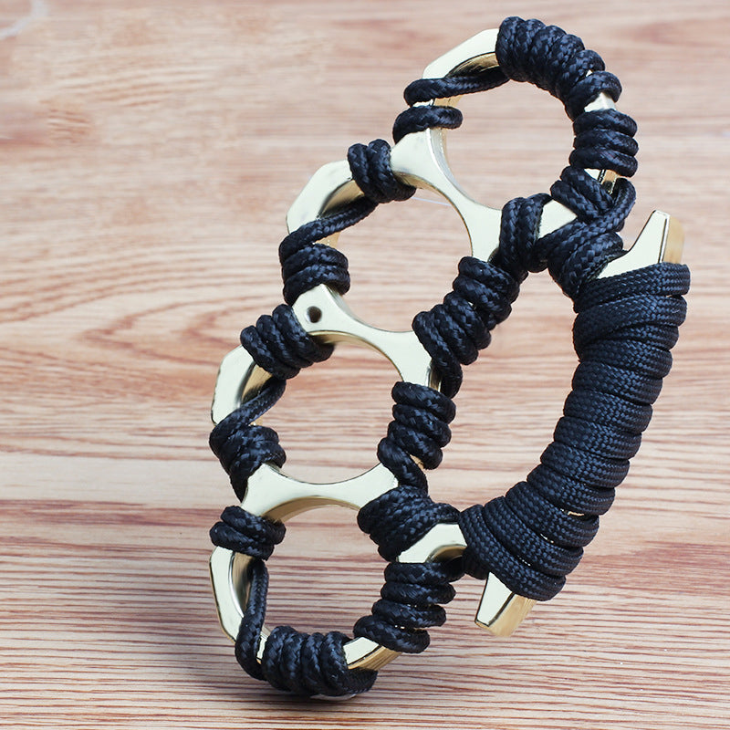 Gold colourway of the woven knuckle duster showing black umbrella rope knotted around the four openings and wrapped palm base on wood