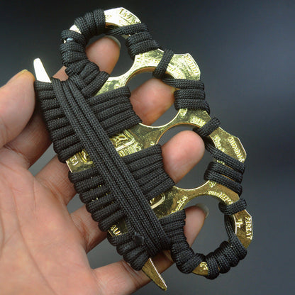 A hand holds a golden zinc alloy knuckle duster wrapped in black paracord, the four finger holes fitted over the fingers and Latin lettering visible on the knuckle faces, against a dark gray background.